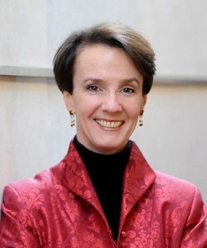Sarah Cleveland smiles directly into camera wearing a short pixie haircut with dangling earrings and a dark turtleneck and a embroidered red blazer